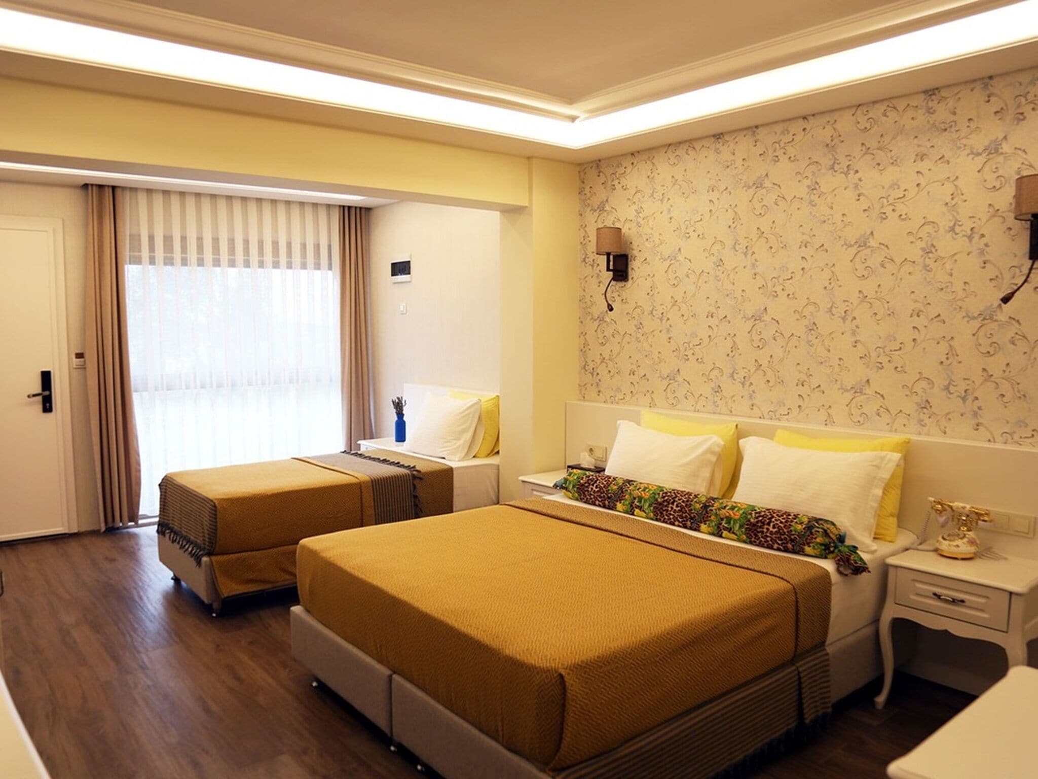 Ein Hotelzimmer mit zwei Betten, einem großen Fenster mit Vorhängen, einer Wandlampe und einem Wandpapier mit Muster.