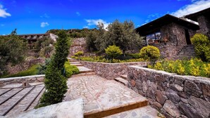 A stone staircase leading to a stone building with a balcony, surrounded by greenery and a clear blue sky.