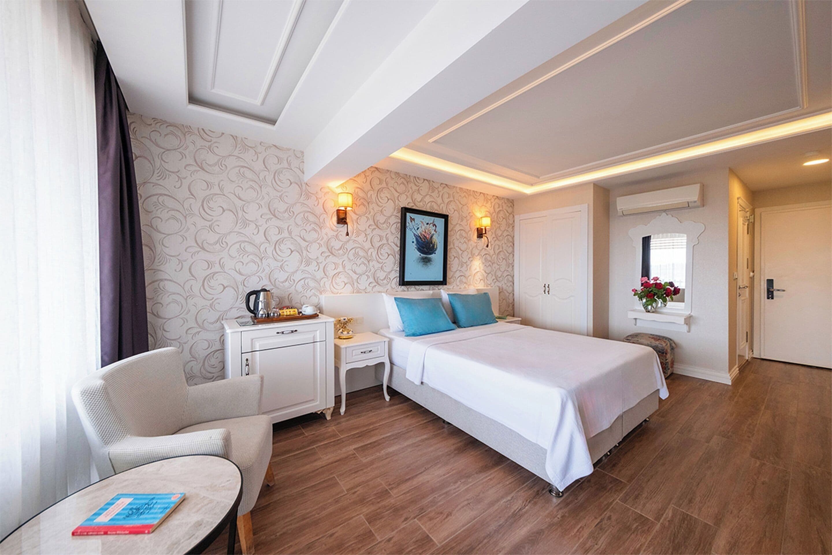 Una habitación de hotel con cama, una silla, una mesita, una mesita de noche, un cuadro en la pared y una ventana con flores.