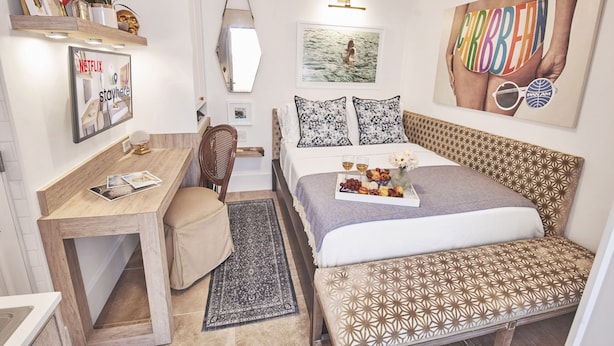 Una habitación de hotel compacta con cama, escritorio y silla. Se observa una repisa en la pared con adornos y un cuadro enmarcado.