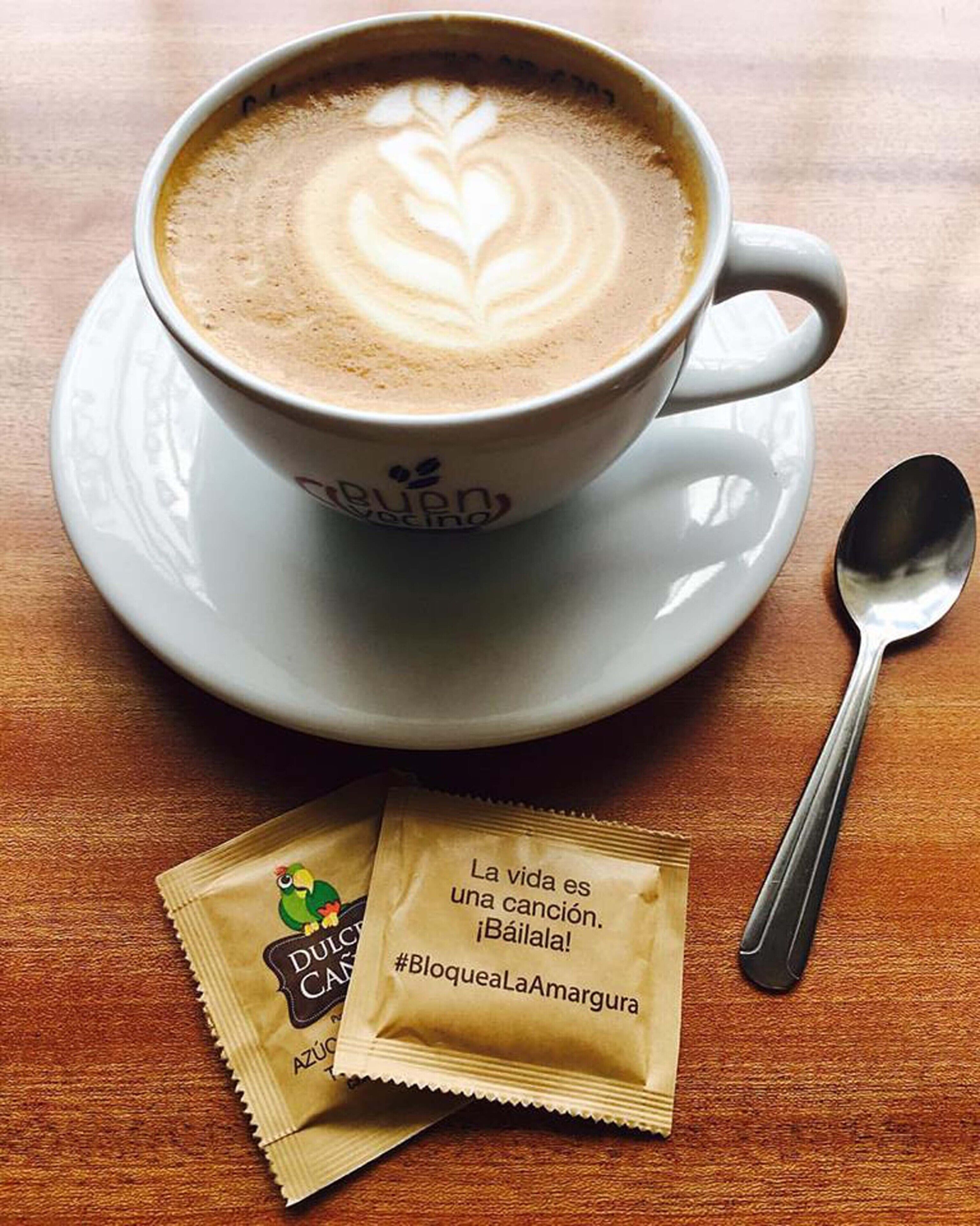 A cup of coffee with latte art on a saucer, a spoon, and two packets of Dulce de Leche Amargura.