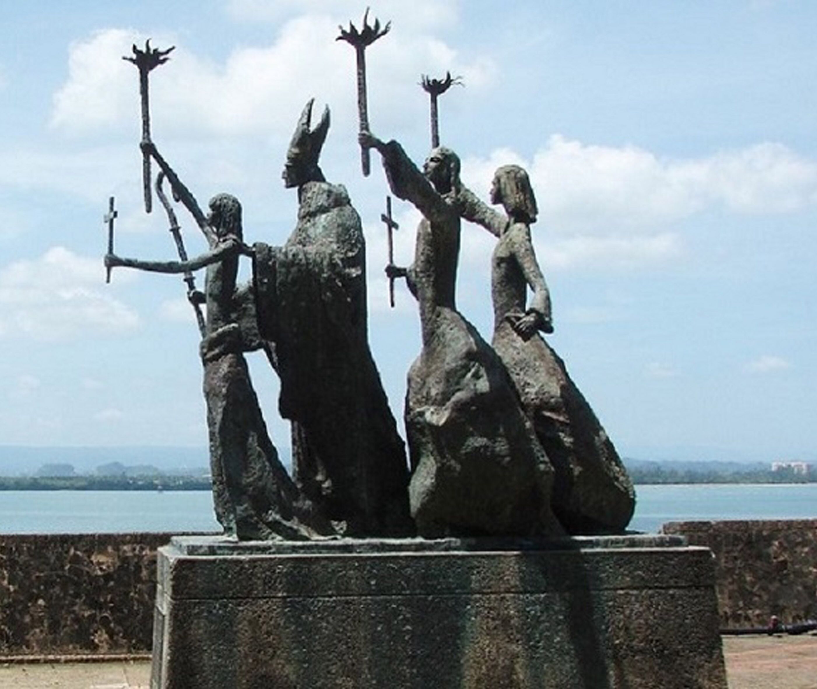 A statue of three figures on a stone pedestal by a waterfront.