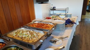 A buffet setup with various dishes, including a large pizza and a casserole, in a modern kitchen area with a wooden wall and a television above the counter. - Hotel Restaurant Teichaue (Adendorf)
