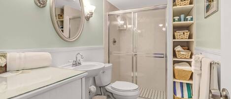 A bathroom with a glass shower enclosure, a white sink, and a toilet. There are wicker baskets on shelves and a mirror with a decorative frame.