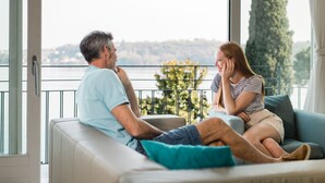 Ein Paar sitzt auf einer Couch vor einem großen Fenster mit Blick auf ein Gewässer und viel Grün.