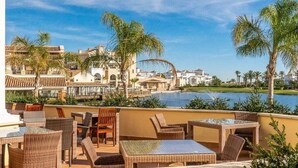 Una terraza en la azotea con mobiliario de mimbre, vista a un lago y edificios de estilo arquitectónico mediterráneo.