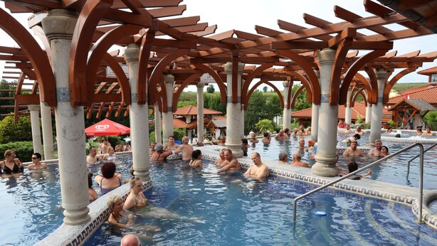 A large outdoor swimming pool with multiple people enjoying the water, surrounded by wooden pergolas and columns.