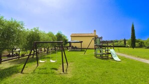 A playground with swings and a slide, a building with a tiled roof, and a grassy area with trees. - PODERE MARCIGLIANO - CORITO (Cortona)
