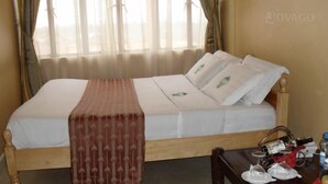 A hotel room with a wooden bed, white linens, a red patterned bedspread, and a window with sheer curtains. - Batian Grand Hotel (Nyeri)