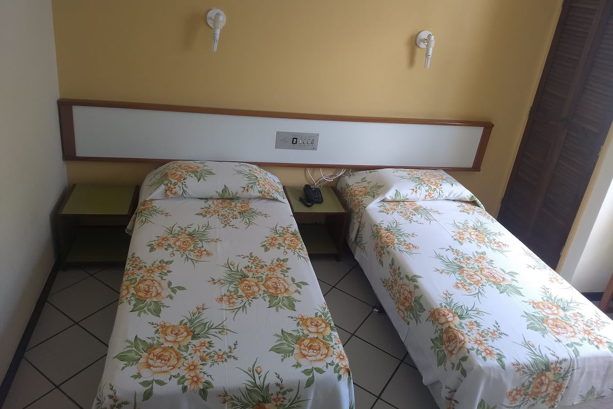 Una habitación de hotel con dos camas, cada una con ropa de cama floral, un cabecero de madera, una mesita de noche y un enchufe en la pared.