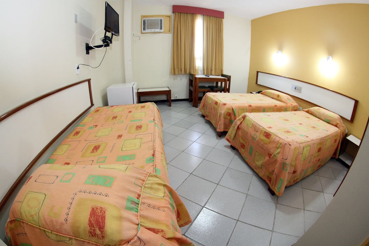 Habitación de hotel con dos camas individuales, un escritorio, una silla, una mesita y un televisor montado en la pared, además de una ventana con cortinas.