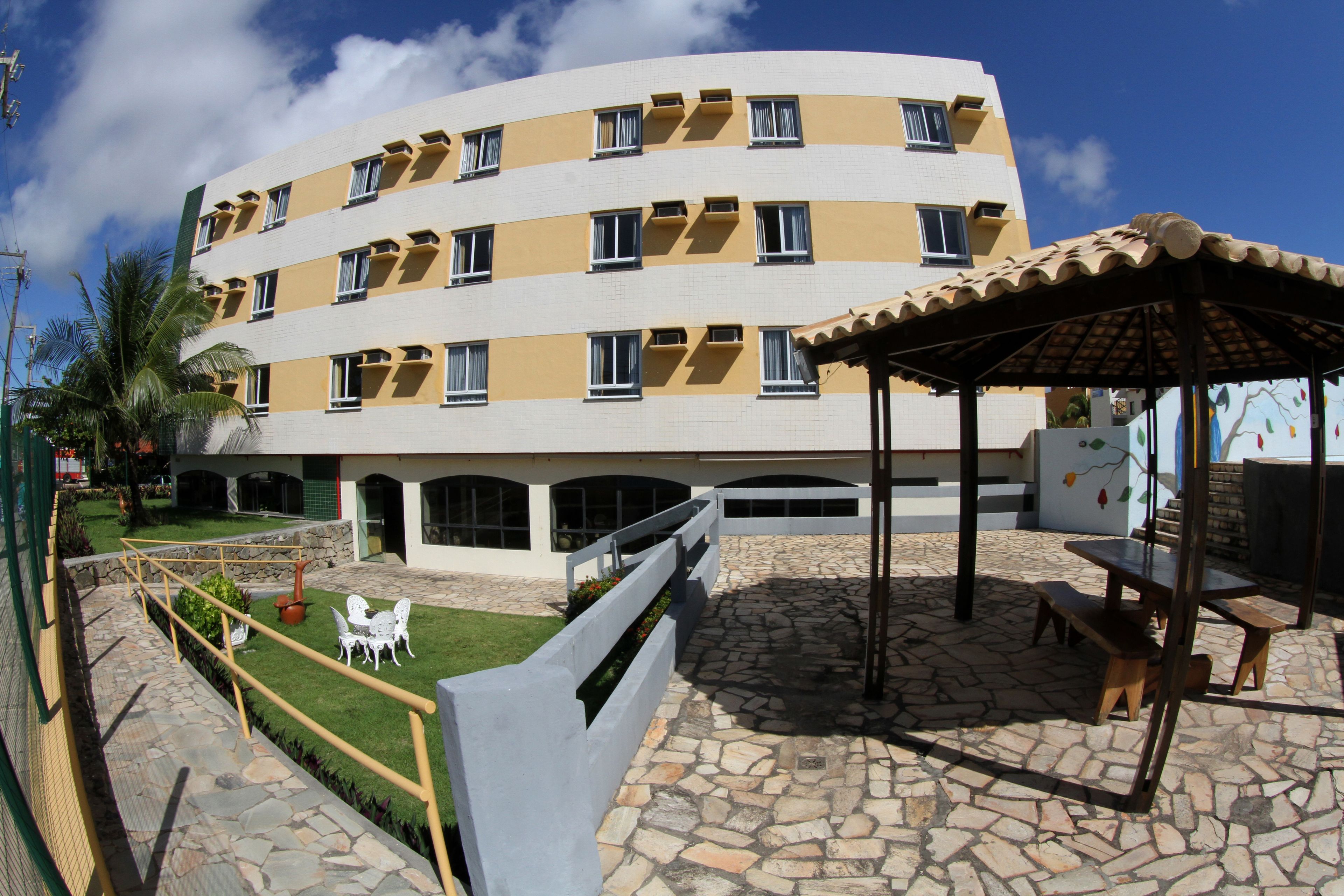 Un immeuble à plusieurs étages doté d’une façade incurvée, d’un espace extérieur aménagé avec des tables et des chaises, et d’une terrasse pavée de pierres.