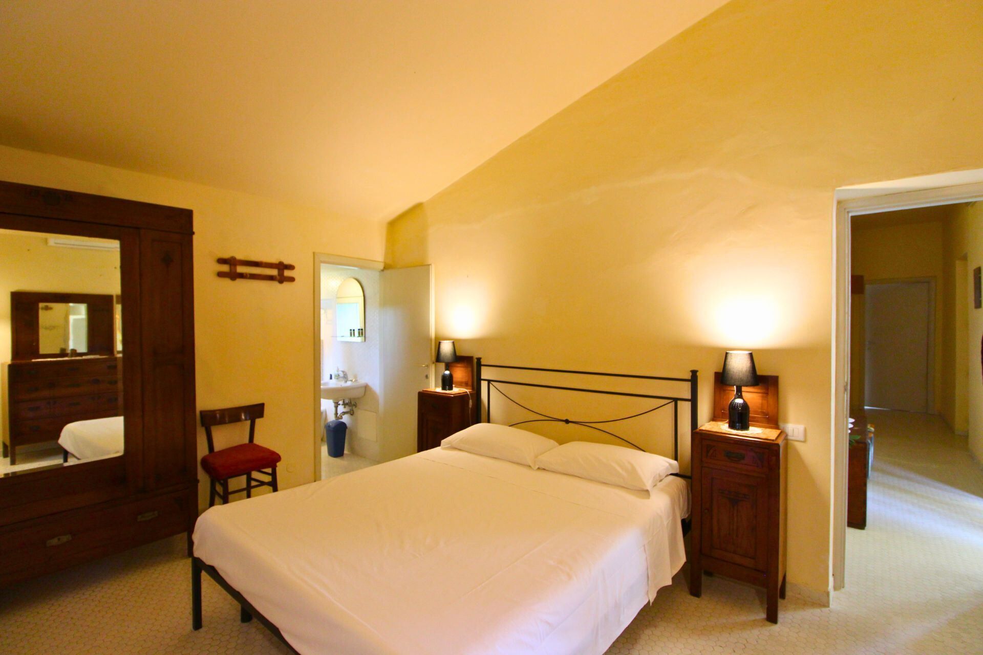 Une chambre à coucher avec un grand lit, deux tables de chevet avec des lampes, une commode en bois, un fauteuil rouge et un miroir.