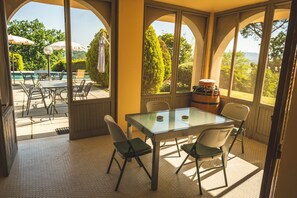Un patio iluminado por el sol con una mesa y sillas de vidrio, con vistas a una piscina y abundante vegetación.