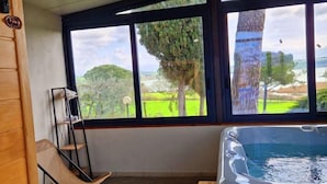 A room with a hot tub, a wooden chair, a small table, and a view of a green landscape through large windows.