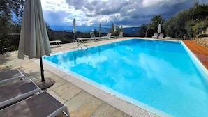 Une piscine rectangulaire à l’eau d’un bleu limpide, entourée de chaises longues et de parasols, avec en arrière-plan des arbres et un ciel nuageux.
