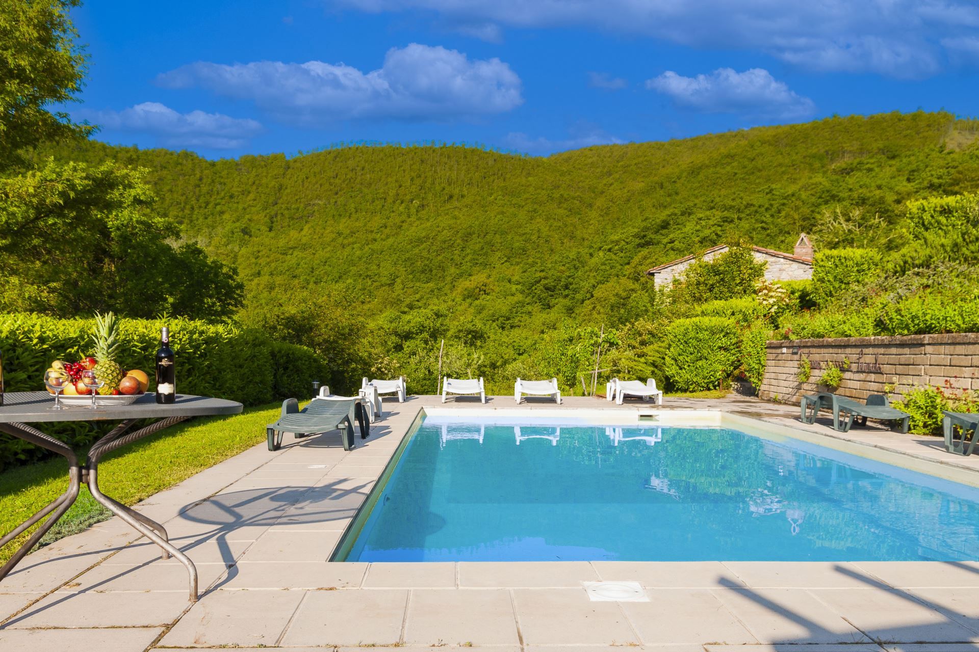 A swimming pool with lounge chairs, a table with fruit and a bottle, and a house in a hilly landscape.