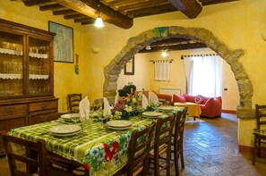 A dining area with a table set for four, a wooden cabinet with glass doors, and a stone archway leading to a living room with a red sofa.
