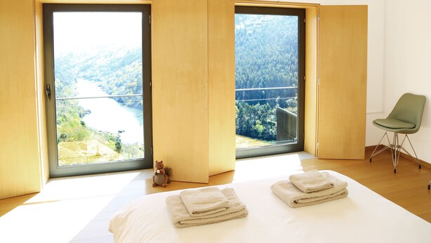 A bedroom with a large bed, two windows with a view of a forest and a bridge, a green chair, and a teddy bear on the floor. - Private Pool with fantastic views over Douro River - Porto & Airport 1H (Marco De Canaveses)