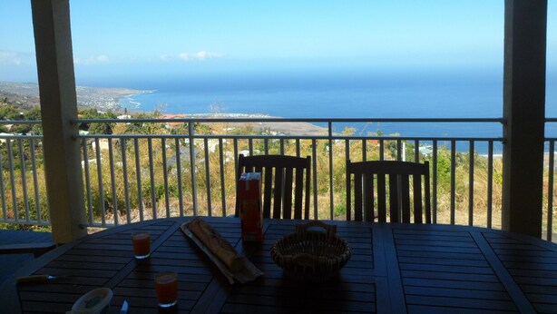 A wooden table with a glass of juice, a baguette, and a basket on a balcony overlooking the ocean.