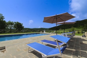 Una piscina con lettini e ombrelloni, circondata da un solarium in pietra e vista sulle colline.