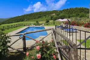 Une piscine entourée d’une clôture en bois, bordée d’un jardin où poussent des rosiers roses, et offrant une vue sur une colline verdoyante.