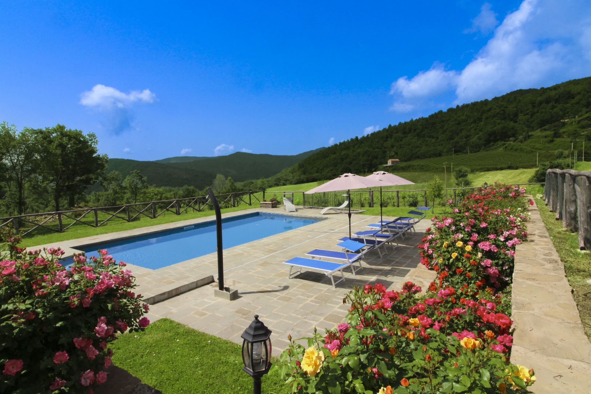 A swimming pool with lounge chairs and a pergola, surrounded by flower beds and a view of hills.
