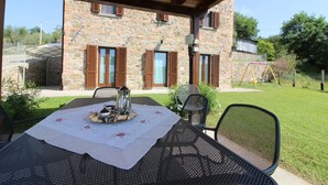 Un espace extérieur aménagé pour les repas, avec une table dressée pour deux personnes, un bâtiment en pierre avec des volets en bois en arrière-plan et une pelouse verdoyante.