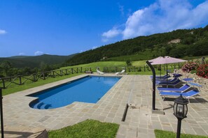 Piscina retangular com deck de pedra, rodeada por uma cerca de madeira, e vista para colinas verdes ao fundo.