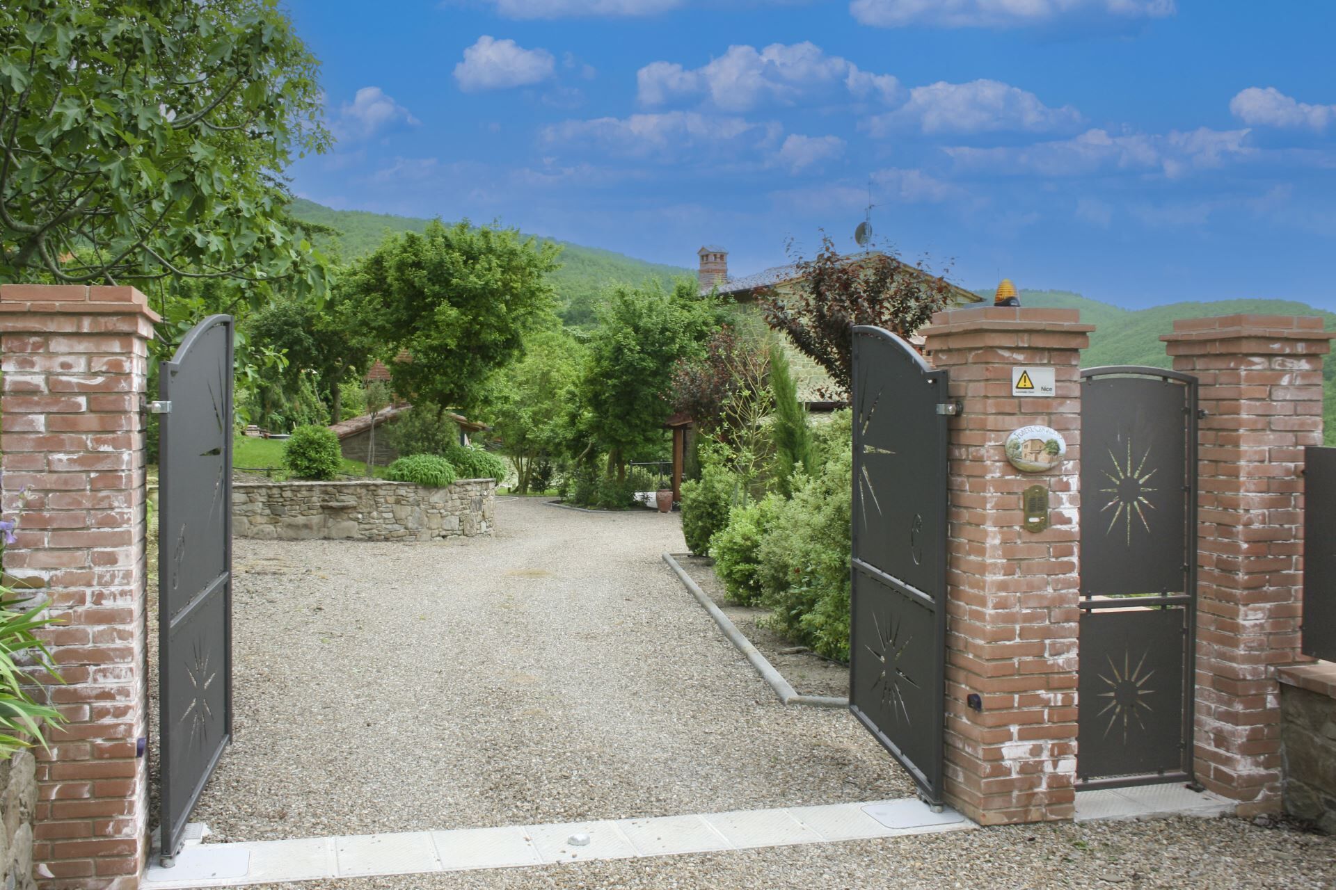 A gated entrance with a gravel path leading to a building, surrounded by greenery and hills.