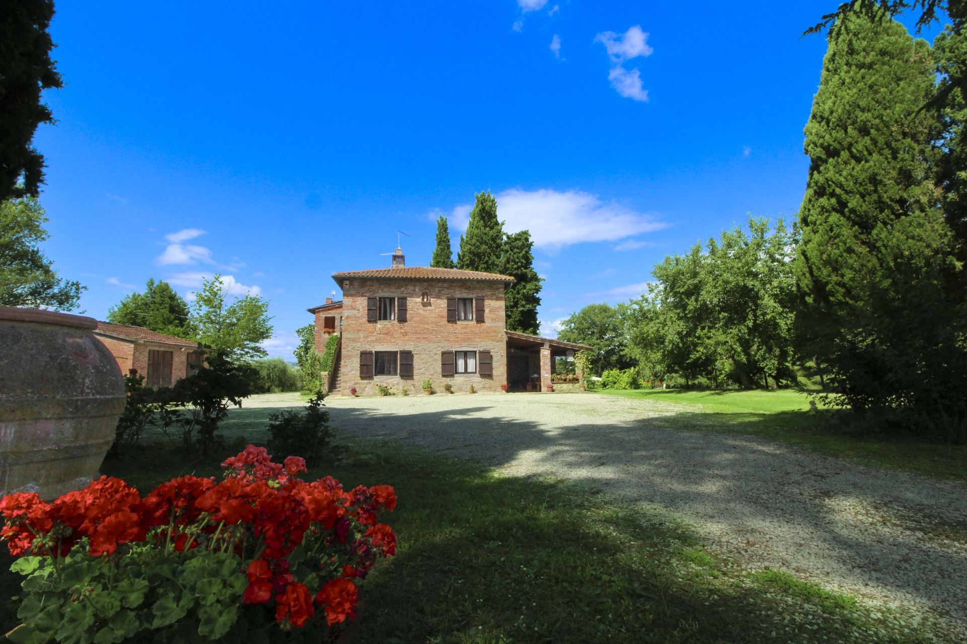 A two-story stone house with a covered porch, surrounded by a gravel driveway and lush greenery.