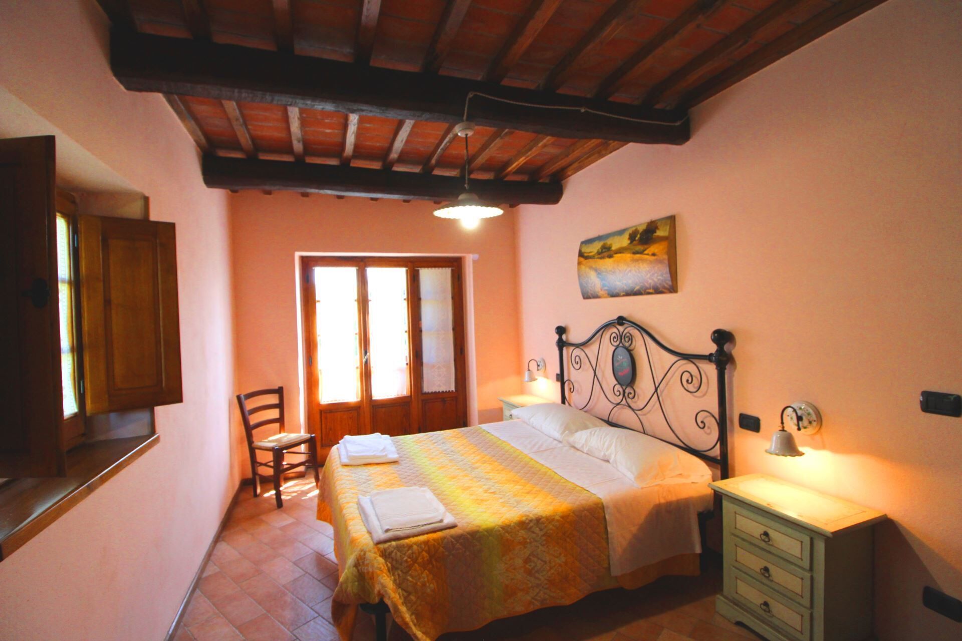 Un dormitorio con cama, una silla, una mesita de noche, un cuadro en la pared y una ventana con cortinas.
