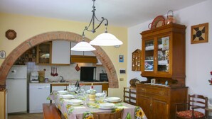 A dining area with a table set for four, a brick archway, a wooden cabinet, and a kitchen in the background.