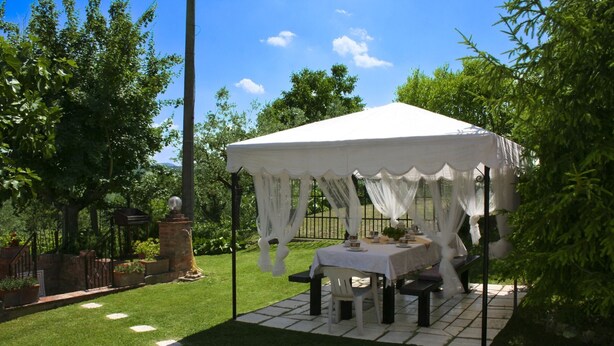 A tented outdoor area with a table set for dining, surrounded by greenery and a brick structure.