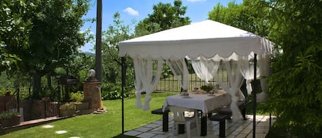 A tented outdoor area with a table set for dining, surrounded by greenery and a brick structure.