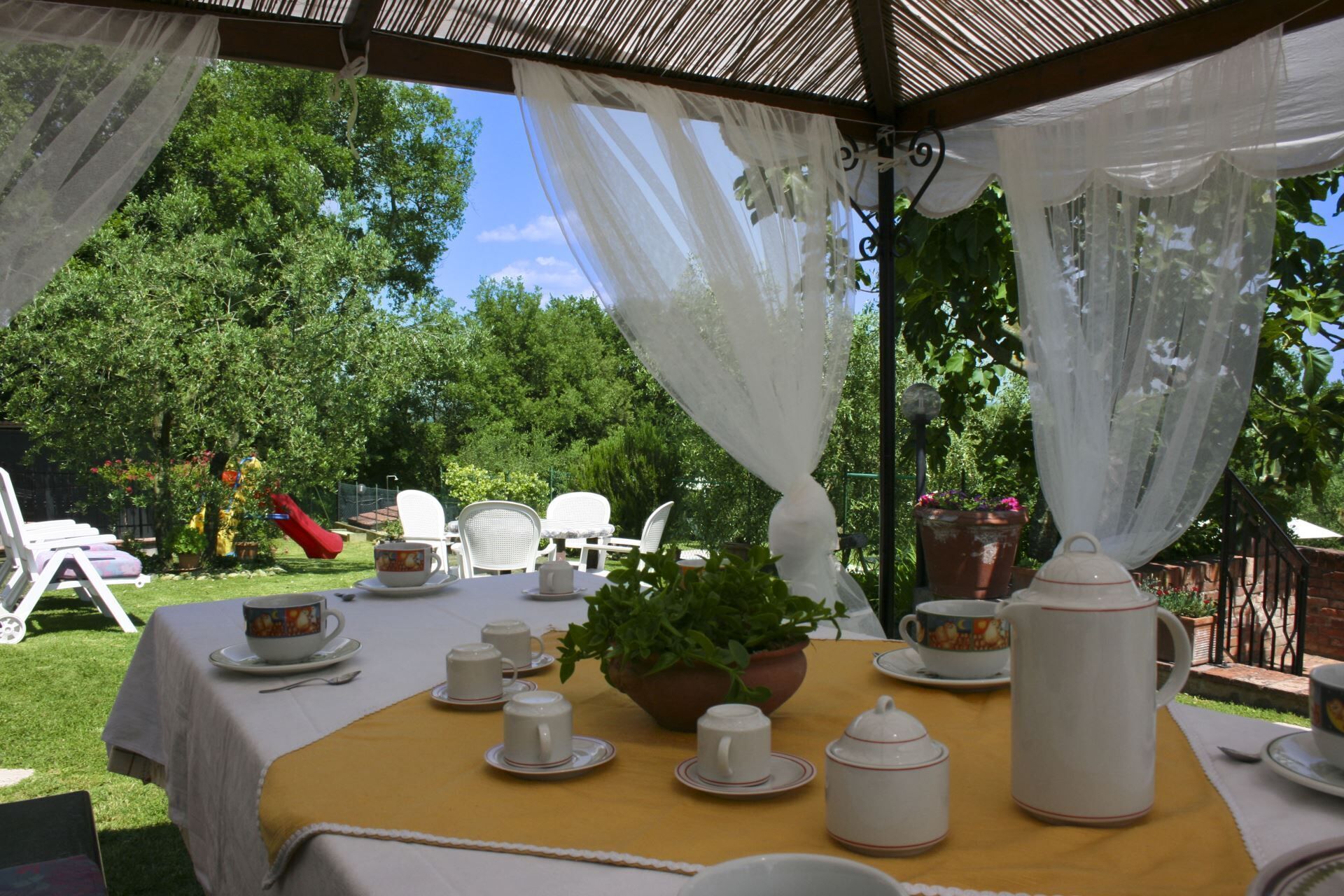 Un área de comedor al aire libre con mesas dispuestas para servicio de té, rodeada de follaje y un cielo despejado.