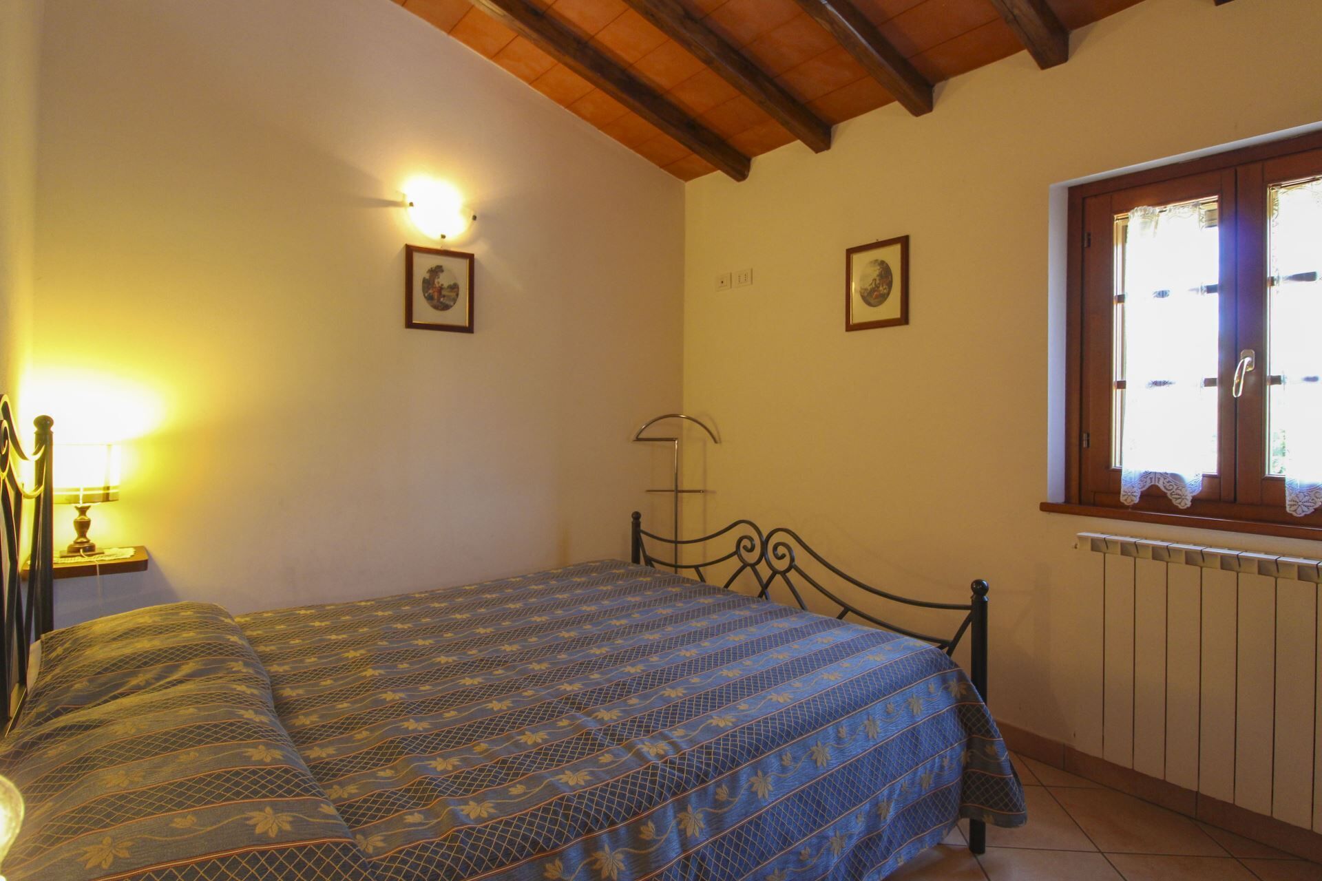 A bedroom with a bed, a nightstand, a lamp, a window with curtains, and two wall-mounted light fixtures.