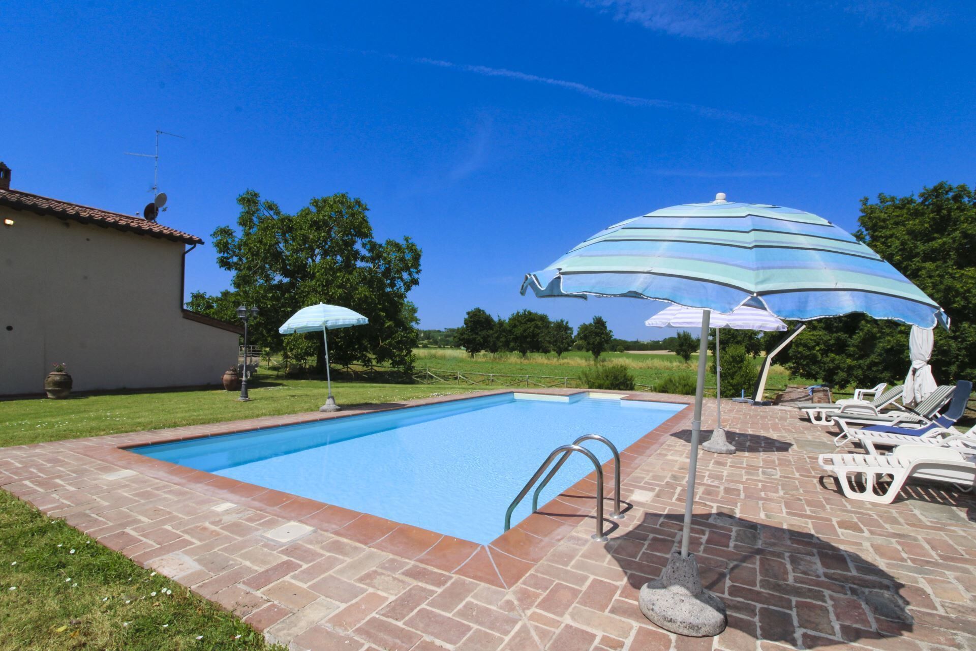 Ein Schwimmbecken mit Ziegelbordstein, zwei Sonnenschirmen, Liegestühlen und einem Gebäude im Hintergrund.