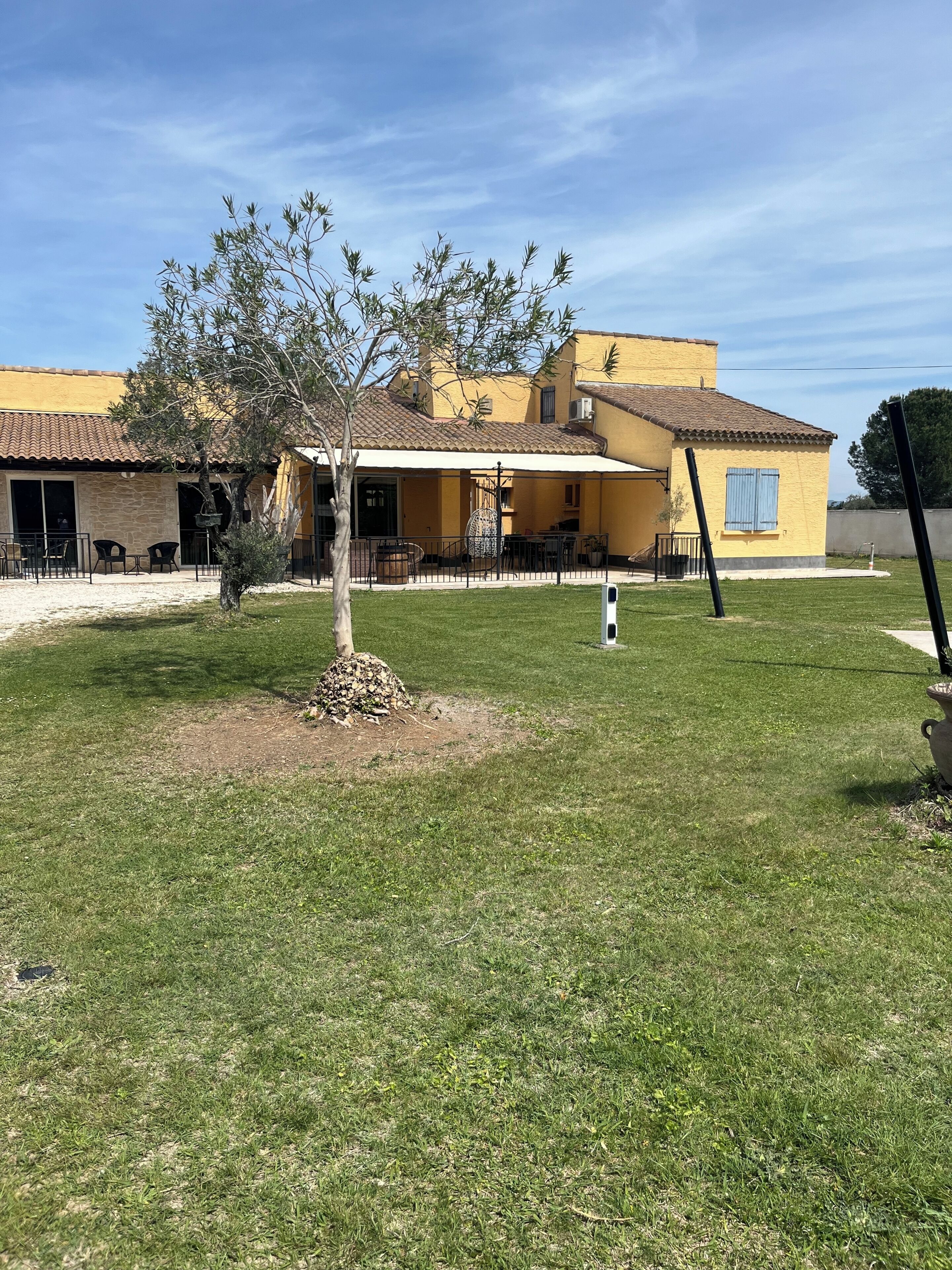 A yellow building with a tiled roof, a covered patio with outdoor furniture, and a small tree in the foreground.