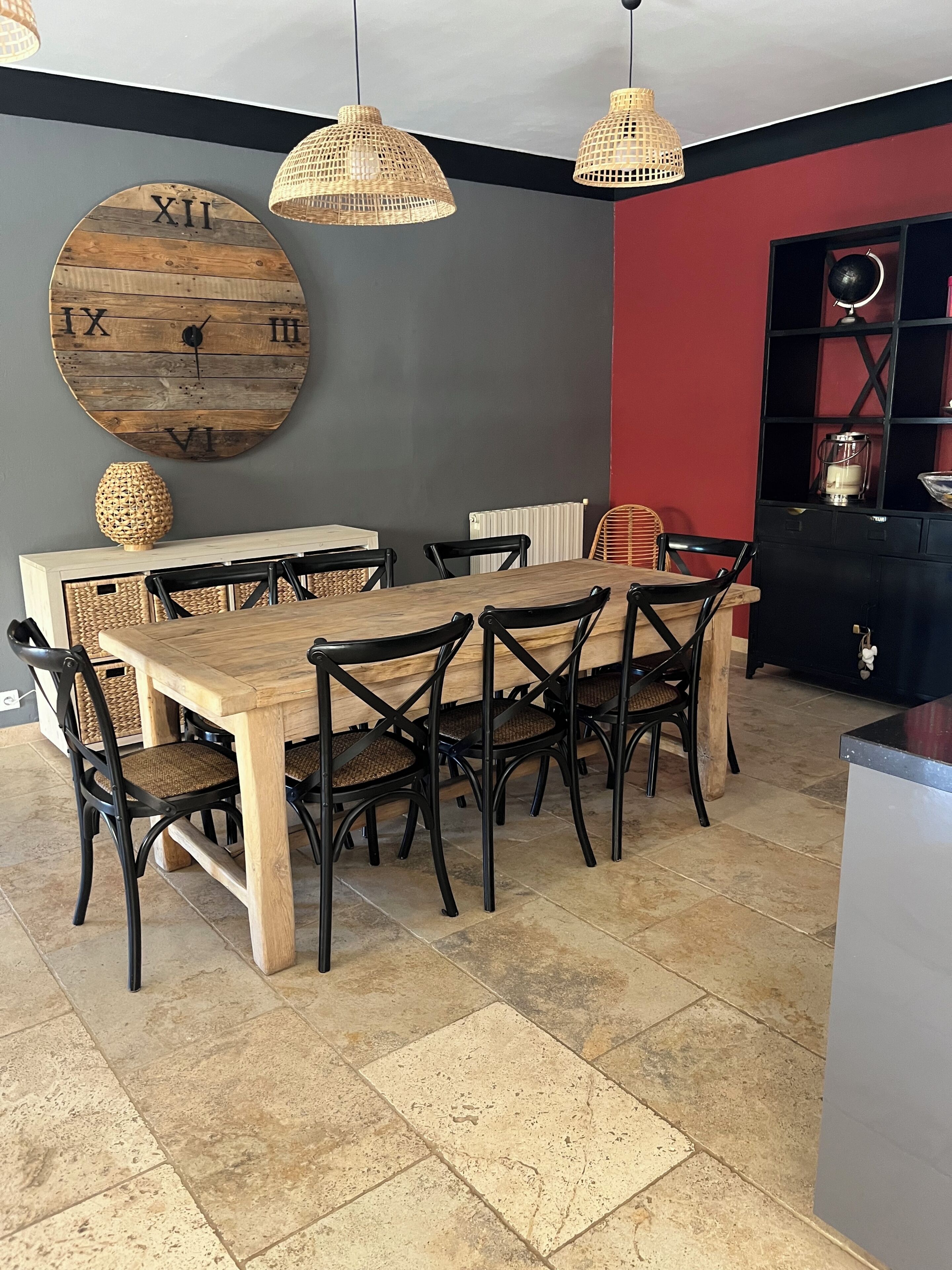 A dining area with a wooden table, black chairs, woven pendant lights, and a large wall clock.