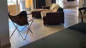 A spacious living area with a sofa, a dining table, a TV mounted on a stone wall, and a large window.