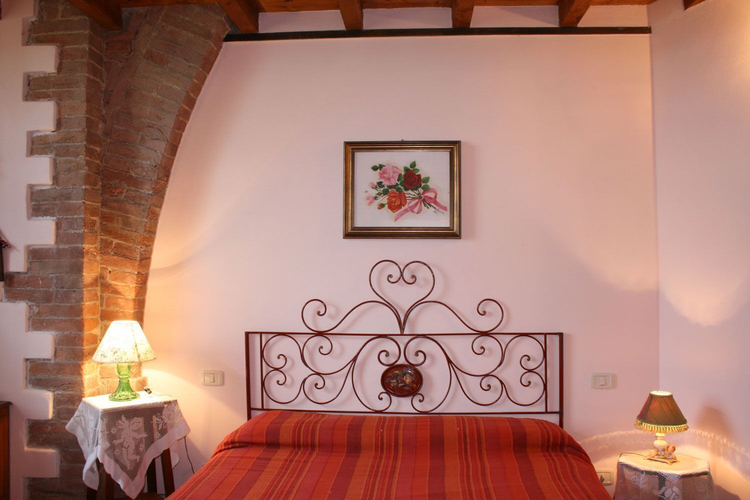 A bedroom with a brick archway, a bed with a red-striped bedspread, a decorative metal headboard, a framed floral artwork on the wall, and two bedside tables with lamps.