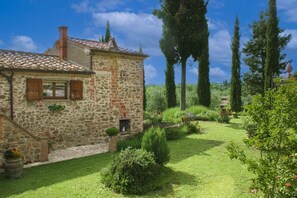 A stone building with a tiled roof, a small garden with various plants, and tall trees in the background.