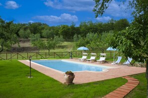 A rectangular swimming pool with a brick deck, surrounded by a low brick wall and a metal fence. There are white lounge chairs with blue umbrellas, and a potted plant in the foreground.