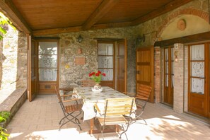 A stone-walled patio with a wooden table, chairs, and a vase of red flowers.