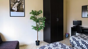 Ein Hotelzimmer mit Bett, Schreibtisch mit Computer, Kleiderschrank, Blumentopf und gerahmtem Foto an der Wand.