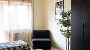 Ein Zimmer mit zwei Betten, einem Sessel, einem Heizkörper, einem Blumentopf und einem Kleiderschrank.