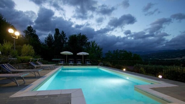 Una piscina con sillas de descanso y sombrillas, rodeada de baldosas de piedra y vistas a árboles y colinas.