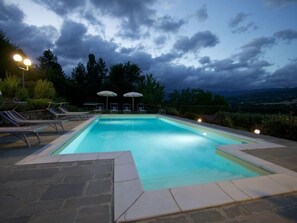 Piscina com espreguiçadeiras e guarda-sóis, rodeada por azulejos de pedra e vista para árvores e colinas.