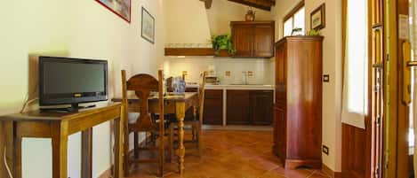Une cuisine avec des meubles en bois, une table à manger avec des chaises, un téléviseur posé sur un meuble et un sol carrelé.